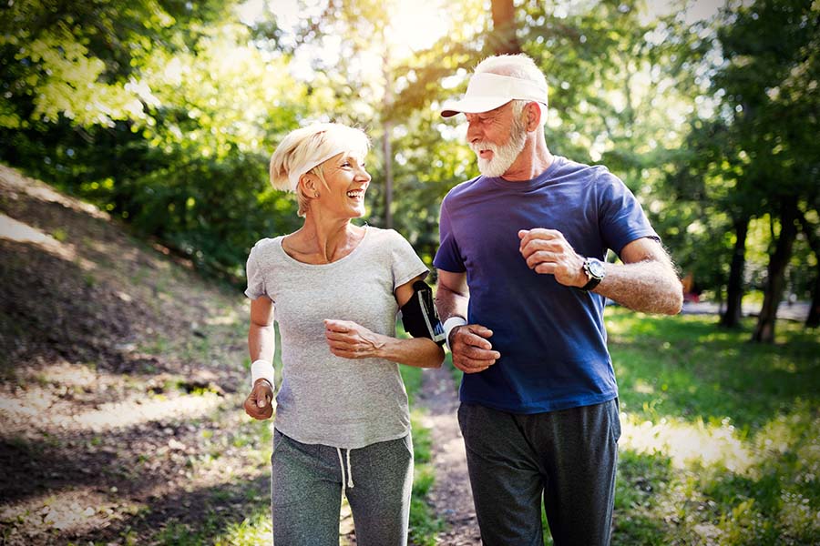 Employee Benefits - Older Couple Smiling While Jogging Through Trail In Forest